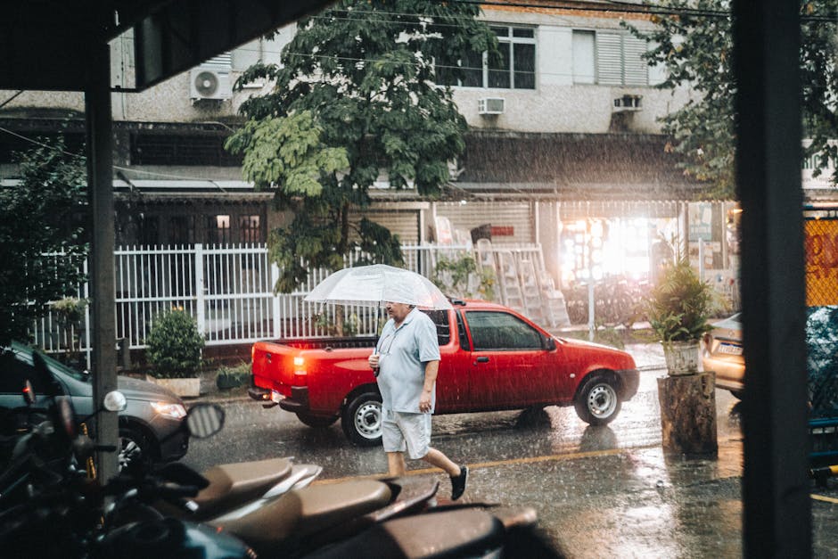 A man holding an umbrella walks by a parked red car during a rainy day in a city setting.