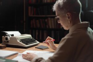 Elderly woman reviewing a manuscript with a typewriter at a wooden desk.