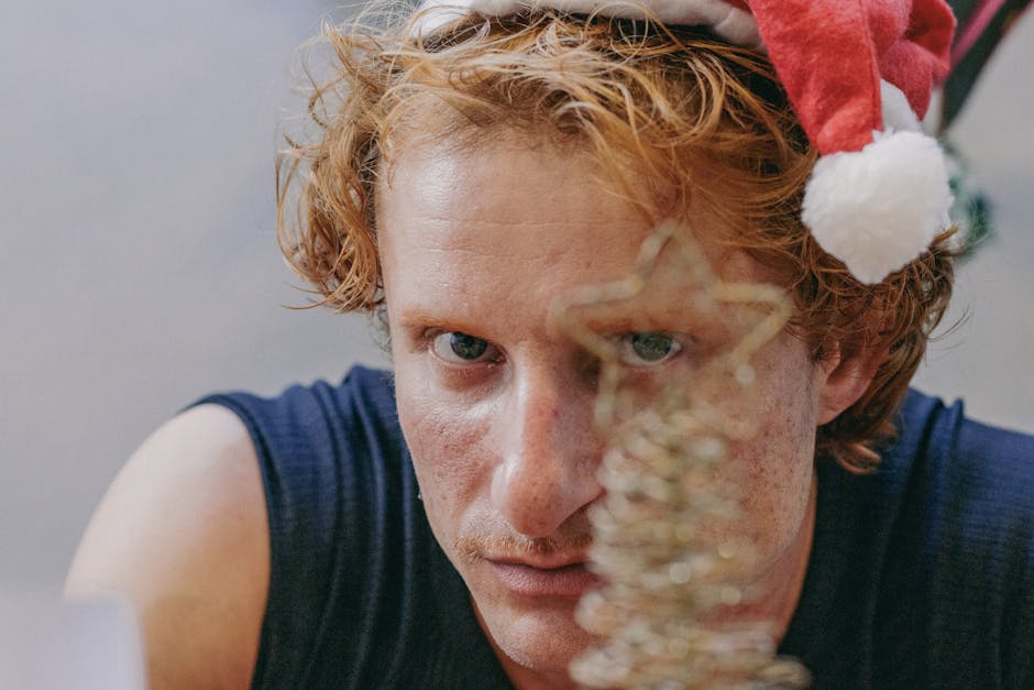 A thoughtful adult man wearing a Santa hat gazes at a shimmering Christmas ornament.