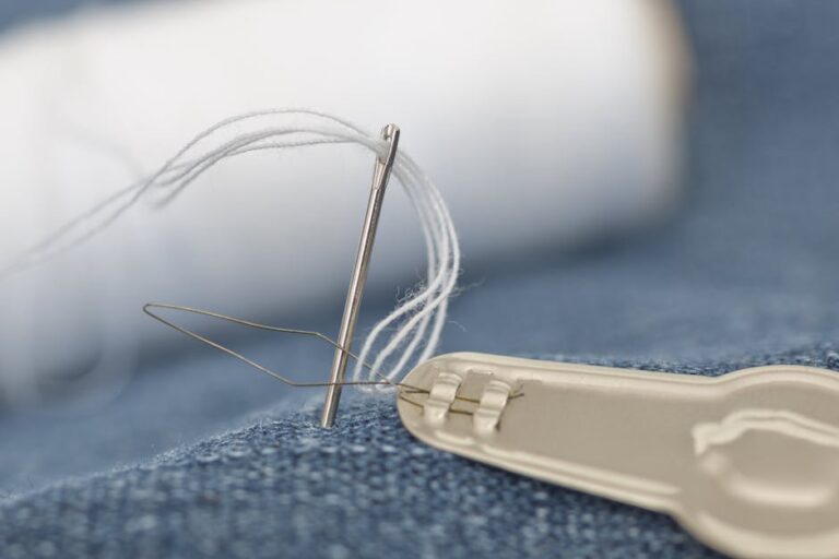 Detailed view of a sewing needle with white thread on blue denim fabric with needle threader.