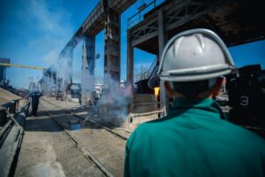 Industrial worker wearing hard hat observing work site with smoke and machinery in a factory setting.
