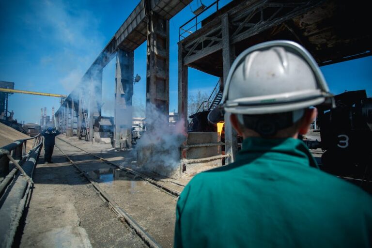 Industrial worker wearing hard hat observing work site with smoke and machinery in a factory setting.