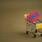 A miniature shopping cart with a bright red 'SALE' tag on a neutral brown background.