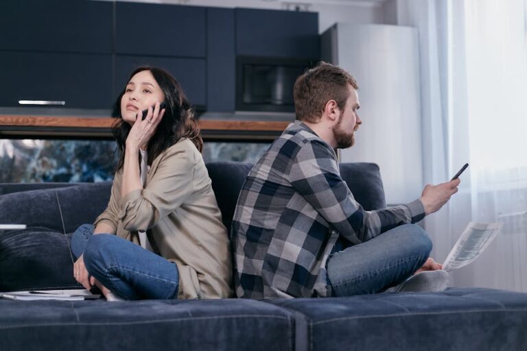 A couple sitting back-to-back on a sofa, expressing financial tension while using phones.