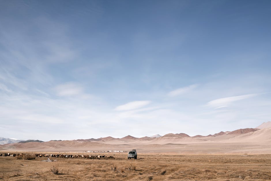 A lone vehicle navigates a remote desert landscape under a vast, clear blue sky.