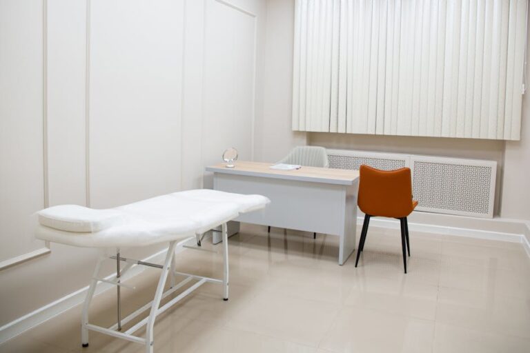 A minimalist medical examination room featuring a desk, chairs, and an examination bed in a sterile, neutral setting.