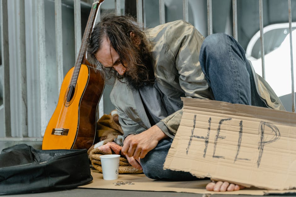 A homeless man sits with a guitar and a help sign on a city street.