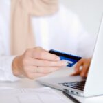 Close-up of a person holding a credit card while using a laptop for online shopping.